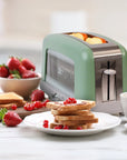A retro-style two-slice toaster in sage green and stainless steel sits on a kitchen counter with toast, strawberries, and a cup nearby, suggesting a breakfast scene.
