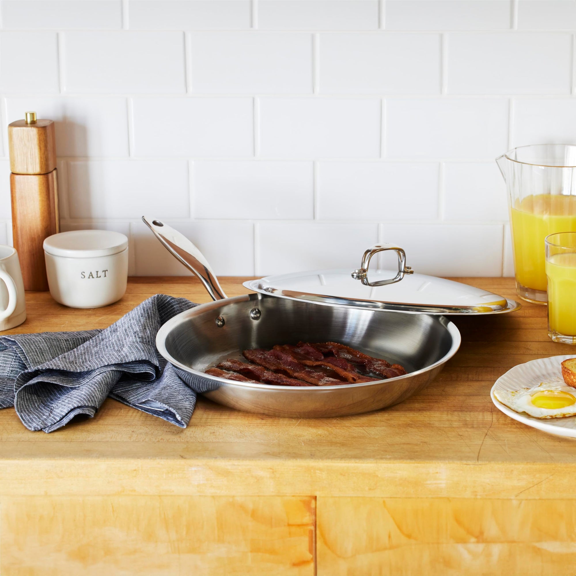 Stainless steel frying pan with lid on a kitchen counter with food and kitchen items.