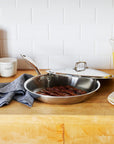 Stainless steel frying pan with lid on a kitchen counter with food and kitchen items.