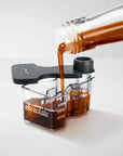 A clear Chocolate attachment with a black lid, shown on a light gray studio background with dark chocolate being poured from a glass bottle into the compartments.