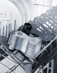A close-up of a dishwasher rack showing a gray and black plastic dish rack insert with a clear water reservoir, alongside white plates and glassware.