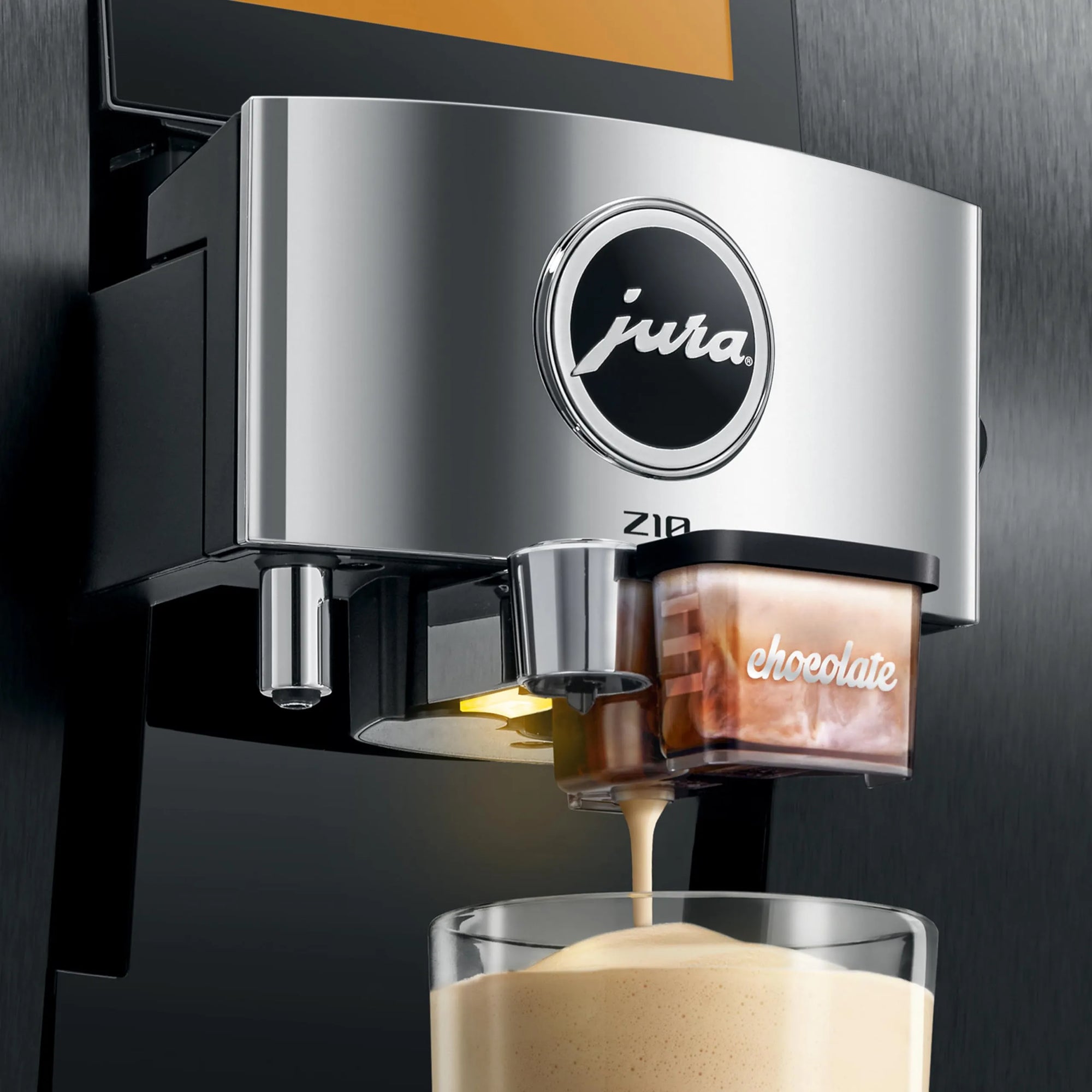 A close-up of a Jura Zib coffee machine dispensing espresso into a glass cup. The machine has a brushed silver metallic front panel with the Jura logo, and a transparent container labeled 'chocolate' sits beside the spout.