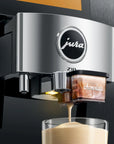 A close-up of a Jura Zib coffee machine dispensing espresso into a glass cup. The machine has a brushed silver metallic front panel with the Jura logo, and a transparent container labeled 'chocolate' sits beside the spout.