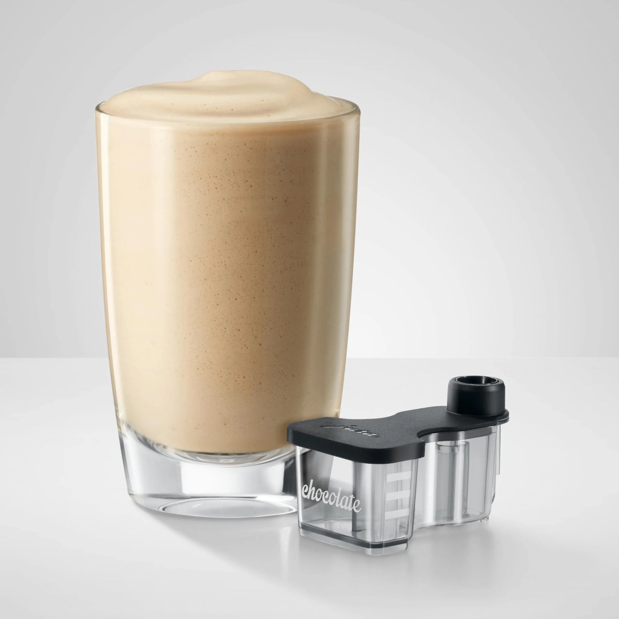 A frothy light brown beverage in a clear glass with a black and clear foam capture device placed beside it on a light gray studio background.