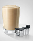 A frothy light brown beverage in a clear glass with a black and clear foam capture device placed beside it on a light gray studio background.