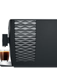 Black coffee machine with wavy design next to a glass of coffee on a white background