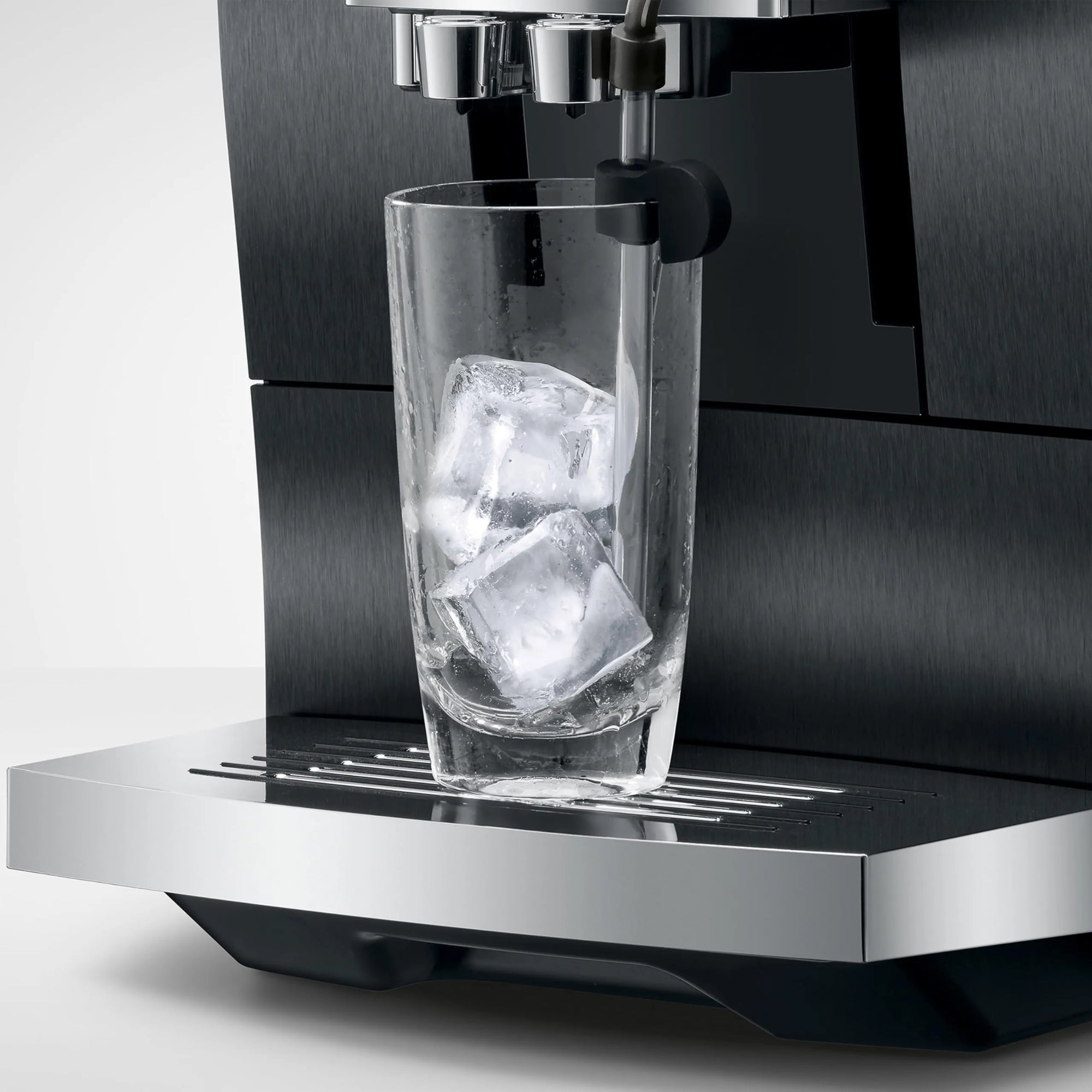 A close-up of a black and stainless steel espresso machine dispensing into a clear glass tumbler filled with ice cubes, set against a clean light gray background.
