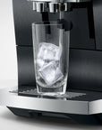 A close-up of a black and stainless steel espresso machine dispensing into a clear glass tumbler filled with ice cubes, set against a clean light gray background.