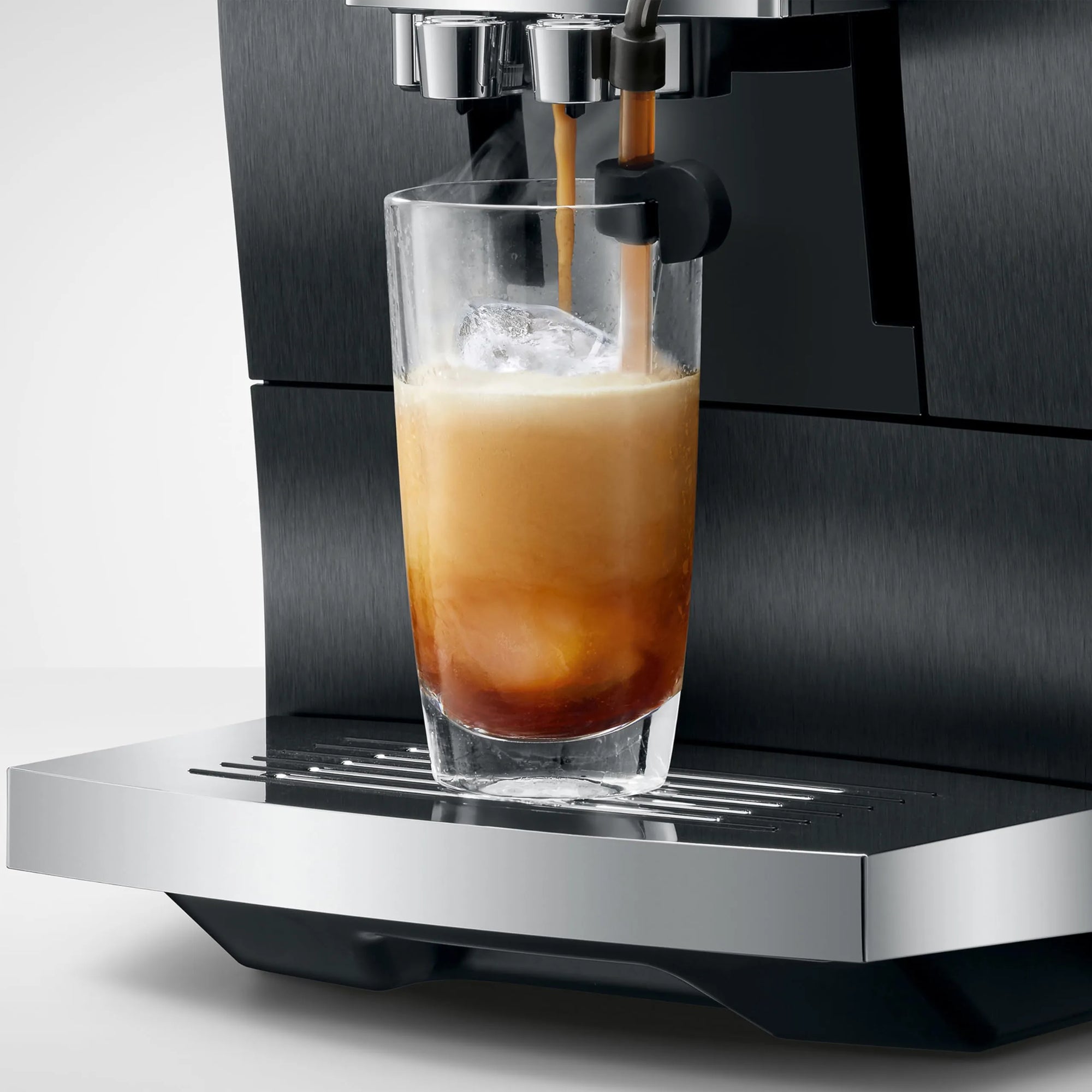 A close-up of a black and stainless steel espresso machine dispensing espresso into a clear glass of iced coffee with ice, set against a clean light gray background.