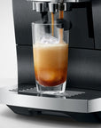 A close-up of a black and stainless steel espresso machine dispensing espresso into a clear glass of iced coffee with ice, set against a clean light gray background.