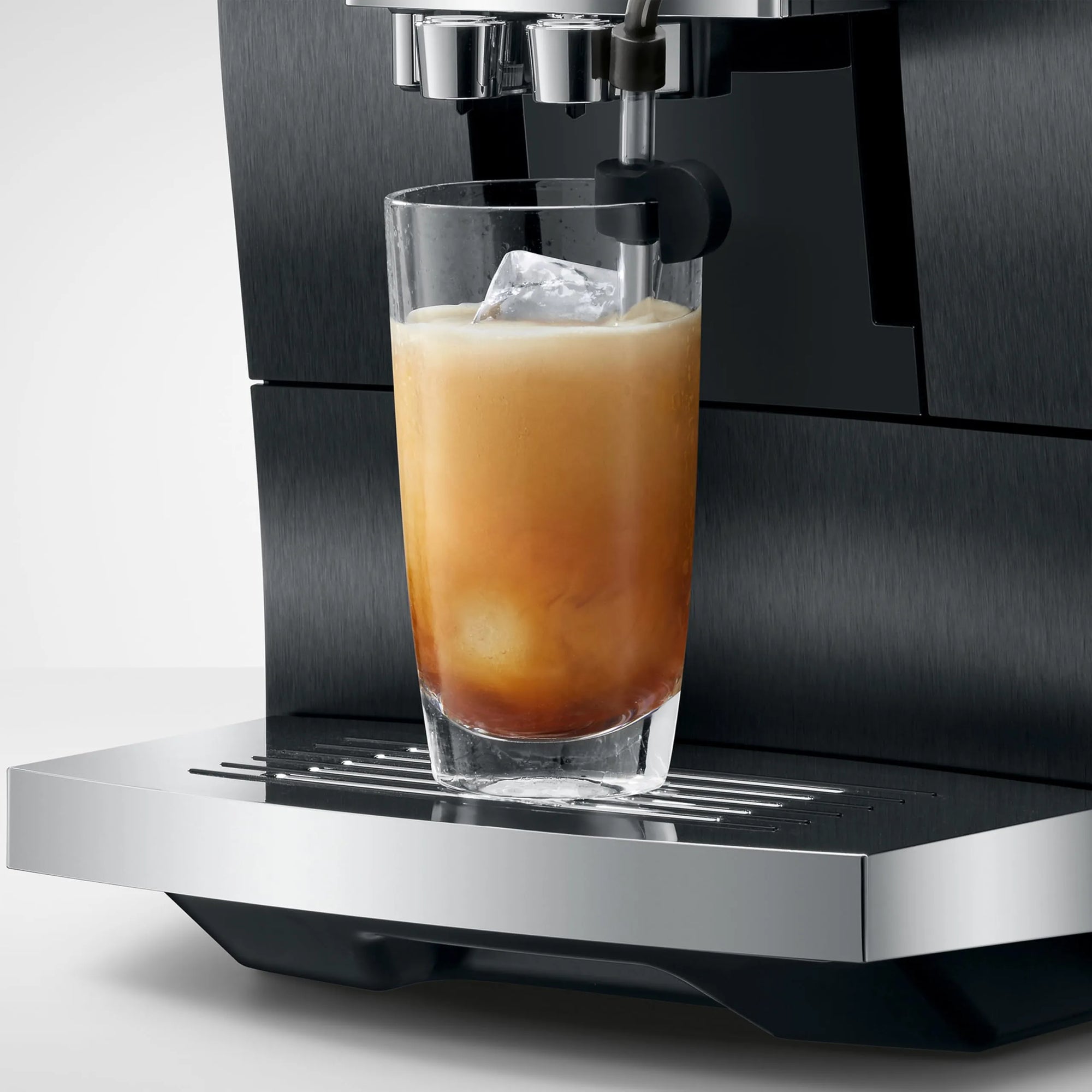 A close-up of a black and stainless steel espresso machine dispensing coffee into a clear glass tumbler with an iced latte, set against a clean light gray background.
