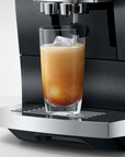 A close-up of a black and stainless steel espresso machine dispensing coffee into a clear glass tumbler with an iced latte, set against a clean light gray background.