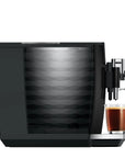 A sleek black coffee machine with a patterned glass water tank and a glass of espresso with foam on its drip tray, set against a solid white background.