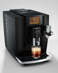 A black Jura coffee machine with a digital display screen, shown dispensing coffee from a glass cup on its drip tray against a light gray background.