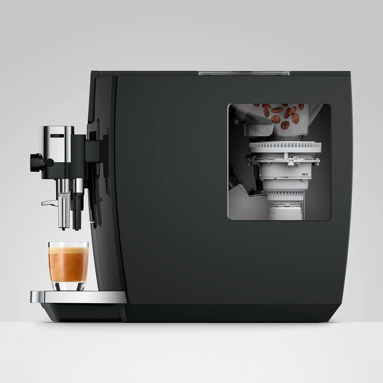 A sleek black coffee machine with a transparent window showing coffee beans and brewing components inside, with a glass of espresso and milk on the drip tray, set against a light gray background.