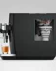 A sleek black coffee machine with a transparent window showing coffee beans and brewing components inside, with a glass of espresso and milk on the drip tray, set against a light gray background.