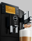 A black Jura E8 automatic coffee machine with a digital display screen, shown dispensing milk foam into a cup of coffee on a light gray background.