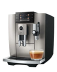 A Jura coffee machine in brushed stainless steel and black, shown with a glass of espresso on the drip tray against a white background.