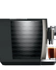 A modern automatic coffee machine in black and silver, shown with water tank and a glass of espresso on its drip tray against a plain white background.