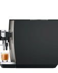 A modern espresso machine with a matte black body and brushed silver metallic accents, shown with a glass of espresso on the drip tray against a plain white background.