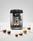 A stainless steel and black espresso machine centered on a light gray studio background, with several glass cups of coffee and milk drinks arranged around it on a white surface.