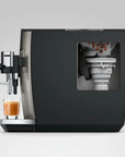 A modern espresso machine in matte black and stainless steel, shown with a glass of espresso on the drip tray and a transparent window revealing the internal bean hopper.