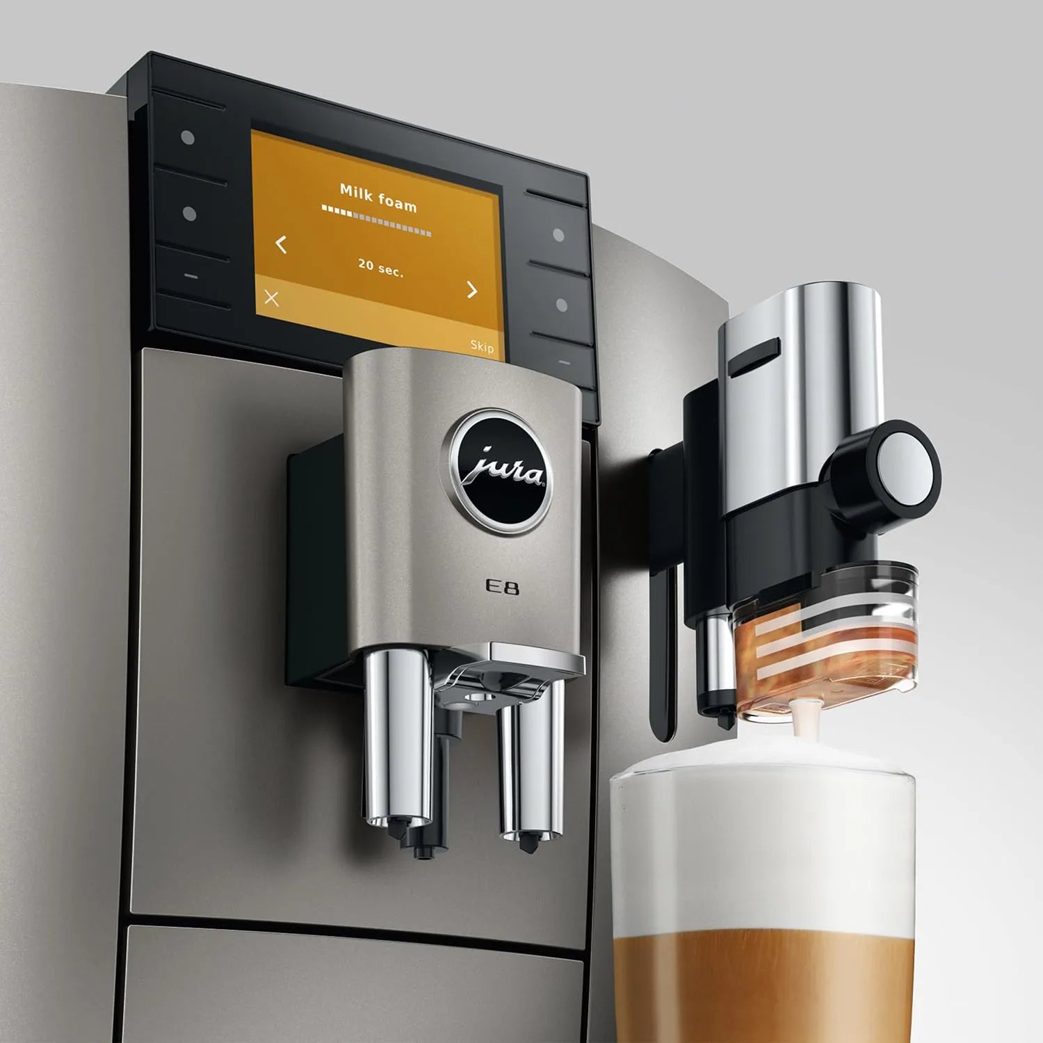 A close-up of a Jura coffee machine dispensing milk foam into a cup of coffee. The machine has a brushed stainless steel body, a black control panel with a yellow-orange display screen, and a milk foam dispenser mounted on the side.