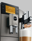 A close-up of a Jura coffee machine dispensing milk foam into a cup of coffee. The machine has a brushed stainless steel body, a black control panel with a yellow-orange display screen, and a milk foam dispenser mounted on the side.