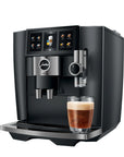 A black and stainless steel Jura coffee machine with a digital touch screen, shown with a glass of espresso and milk on the drip tray against a white background.