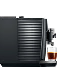 A sleek black coffee machine with a glass water tank and a glass of espresso on its drip tray, set against a solid white background.