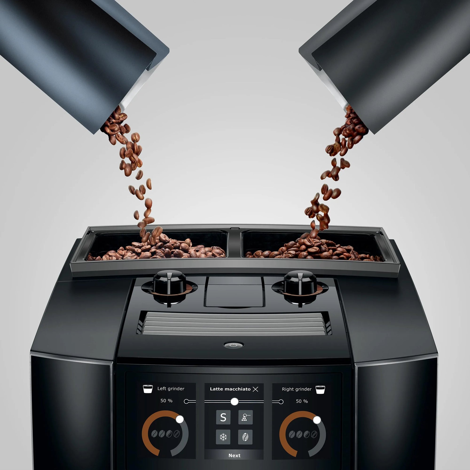 A black coffee machine with two bean hoppers pouring roasted coffee beans into the top compartments, shown on a light gray studio background.
