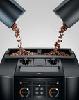 A black coffee machine with two bean hoppers pouring roasted coffee beans into the top compartments, shown on a light gray studio background.