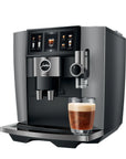 A sleek Jura coffee machine in dark gray and black, featuring a digital touch screen and dispensing spouts, with a glass of espresso and milk foam on the drip tray, set against a plain white background.
