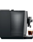 A sleek black and dark gray coffee machine with a ribbed water tank, shown on a plain white background with a small glass of espresso underneath the drip tray.