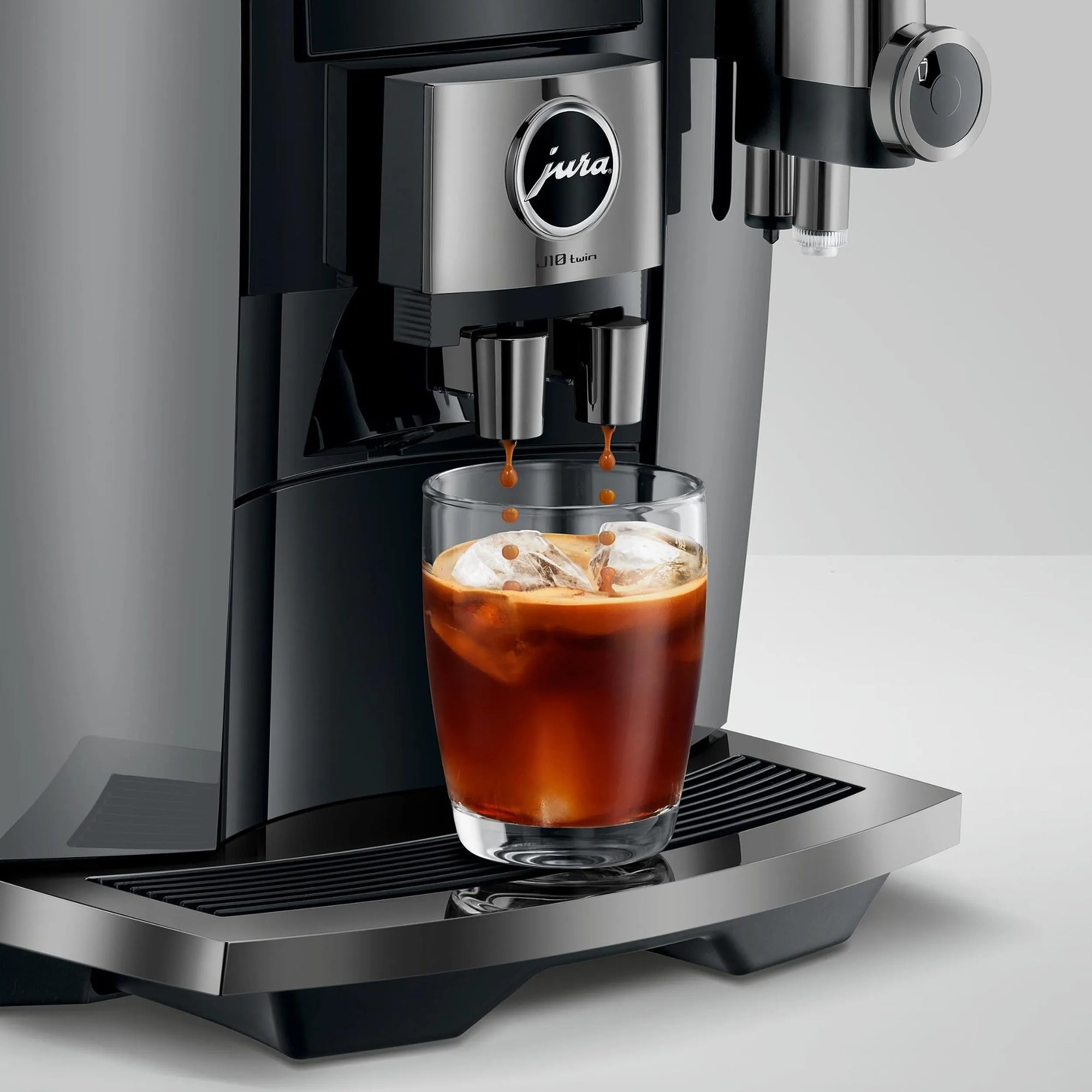 A close-up of a Jura coffee machine dispensing espresso into a clear glass filled with iced coffee, set against a clean light gray studio background.