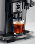 A close-up of a Jura coffee machine dispensing espresso into a clear glass filled with iced coffee, set against a clean light gray studio background.