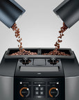 A modern espresso machine with two bean hoppers pouring coffee beans into the hopper compartments, shown against a light gray studio background.