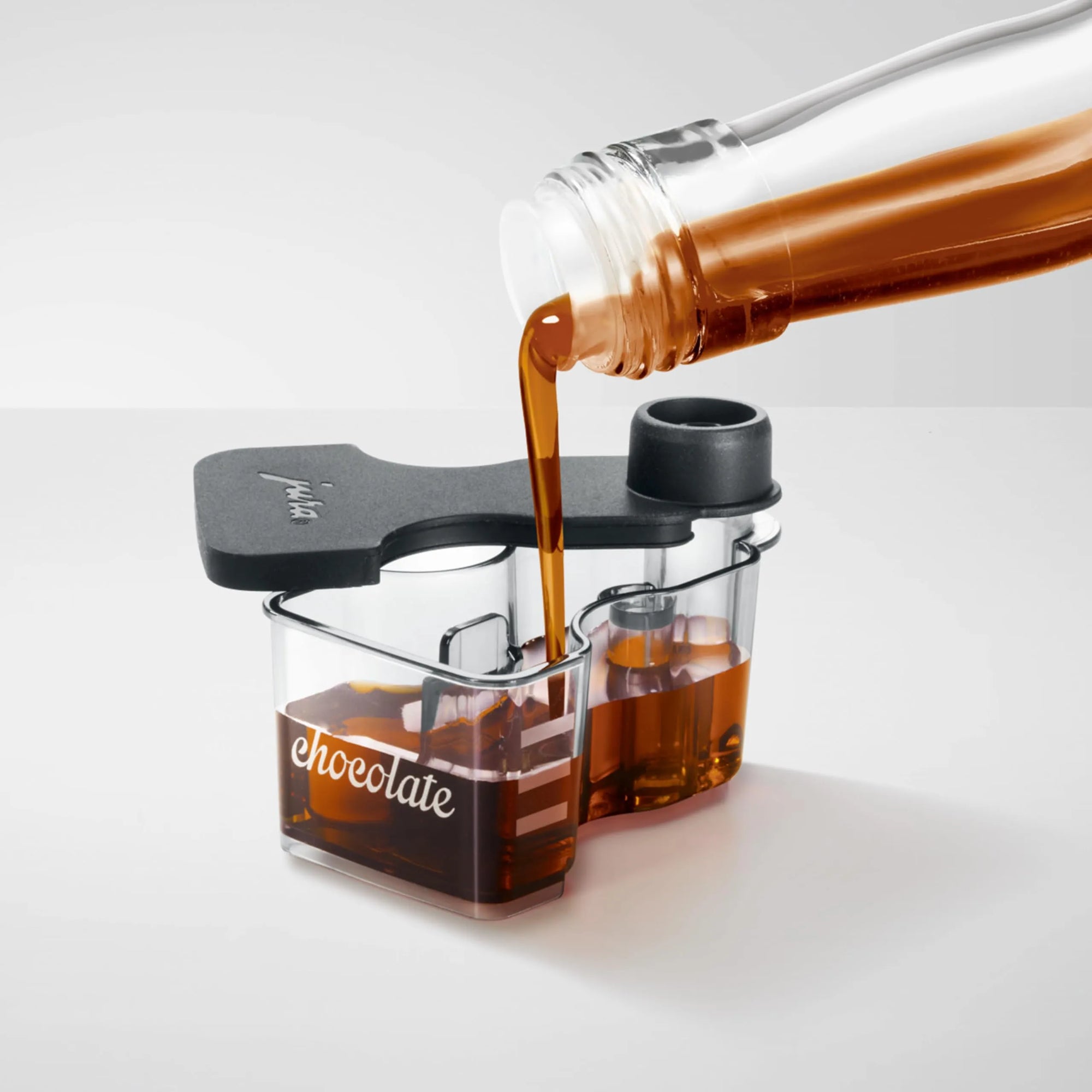 A clear Chocolate attachment with a black lid, shown on a light gray studio background with dark chocolate being poured from a glass bottle into the compartments.