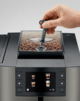 A close-up of a hand inserting a black key into a transparent bean hopper filled with roasted coffee beans on a modern espresso machine.