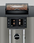 A Jura X10 espresso machine in brushed stainless steel with a black control panel and a transparent bean hopper filled with coffee beans, shown against a light gray background.