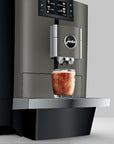A sleek Jura commercial-style coffee machine in dark gray and black, shown dispensing an iced coffee into a clear glass on a metal tray against a light gray background.