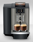 A modern automatic coffee machine in black and stainless steel, shown with two glasses of espresso on the drip tray against a light gray studio background.