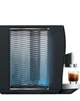A sleek black coffee machine with a textured glass water tank and a small glass cup of espresso on a silver tray, shown against a solid white background.