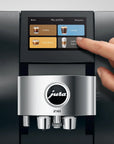A close-up of a Jura Z10 coffee machine with a human hand touching the touchscreen display, set against a dark metallic background.