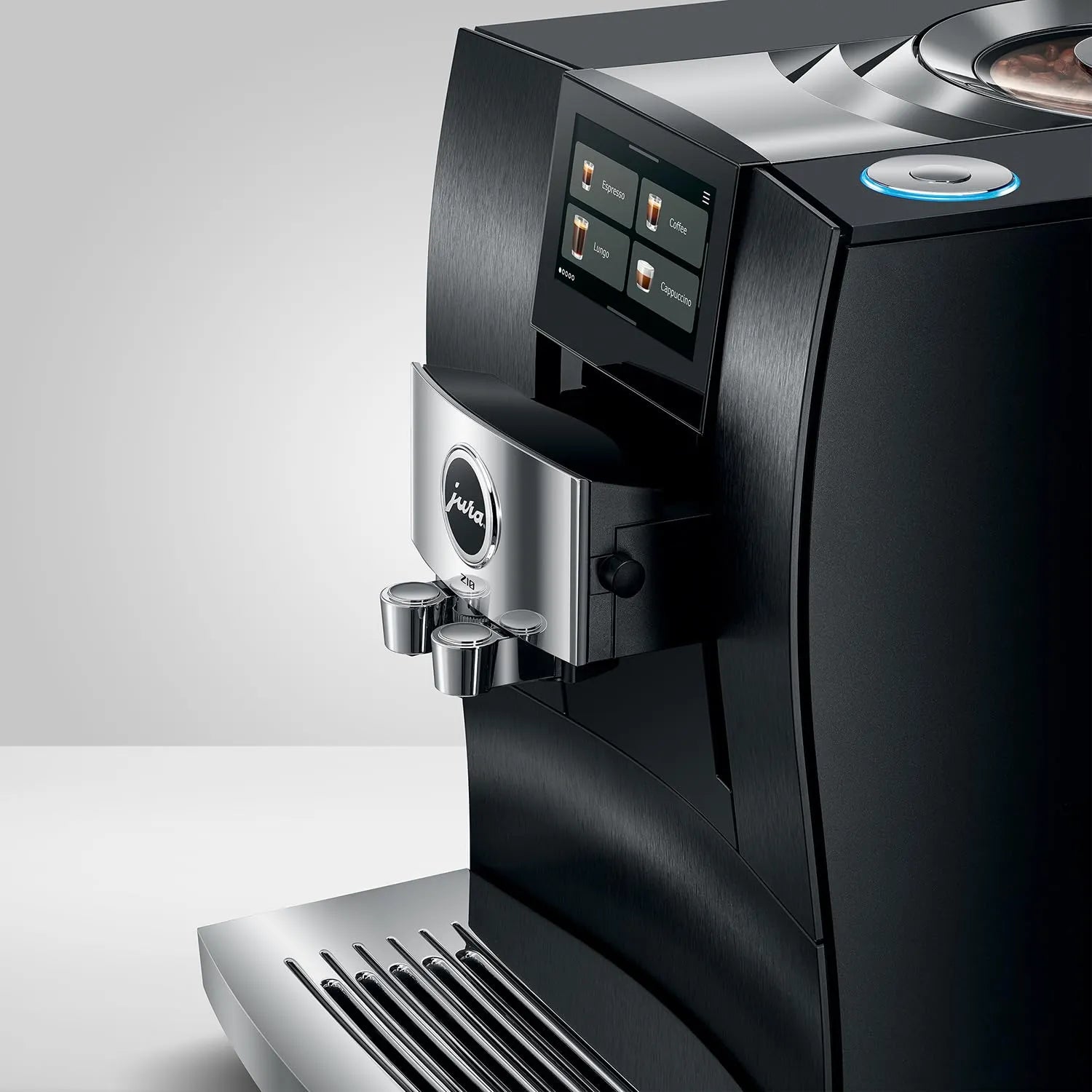A close-up studio shot of a Jura coffee machine in matte black and brushed silver, showing the front display screen, water outlets, drip tray, and part of the bean hopper against a light gray background.