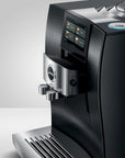 A close-up studio shot of a Jura coffee machine in matte black and brushed silver, showing the front display screen, water outlets, drip tray, and part of the bean hopper against a light gray background.