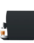 A sleek, modern espresso machine in matte black with a stainless steel drip tray holding a freshly brewed espresso shot, set against a solid white background.