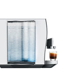 A modern white coffee machine with a textured clear water tank and a small glass of espresso on its drip tray, shown against a solid white background.