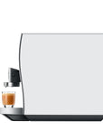 Side view of a modern white coffee machine with a glass of espresso and milk coffee on the drip tray, shown against a solid white background.