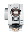 Top view of a modern stainless steel coffee machine with a visible bean hopper filled with coffee beans and a small cup of brewed coffee at the front output tray, shown on a plain white background.